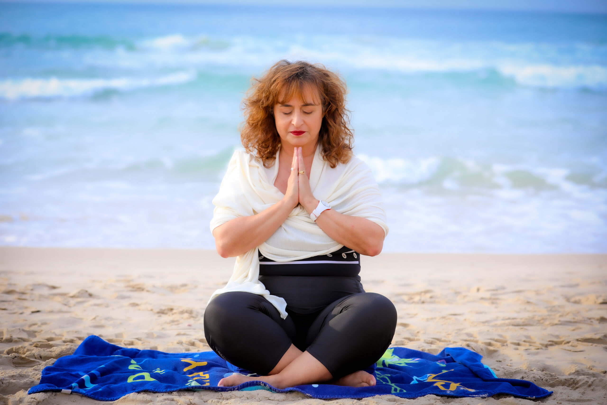 Woman sitting in a lotus pose meditating on a sandy beach in front of breaking waves, wearing dark clothing and an orange wrap.